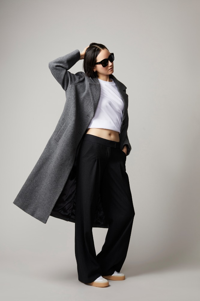 Model wearing a grey wool coat, white crop top, black trousers, and sunglasses against a plain grey backdrop.