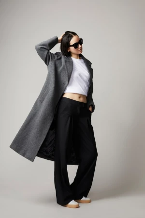 Model wearing a grey wool coat, white crop top, black trousers, and sunglasses against a plain grey backdrop.