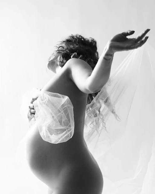 Striking black and white image of a nude pregnant woman holding sheer fabric, with a white background, emphasizing form and texture.