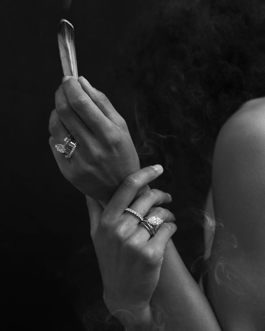 Close-up black and white shot of a person holding sage, adorned with multiple rings, set against a dark background with smoke wisps.