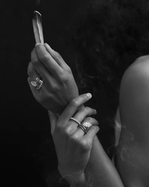 Close-up black and white shot of a person holding sage, adorned with multiple rings, set against a dark background with smoke wisps.