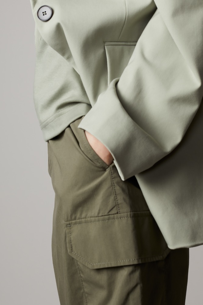 Cropped view of a person in olive green cargo pants and a light green jacket with a large black button against a neutral background.