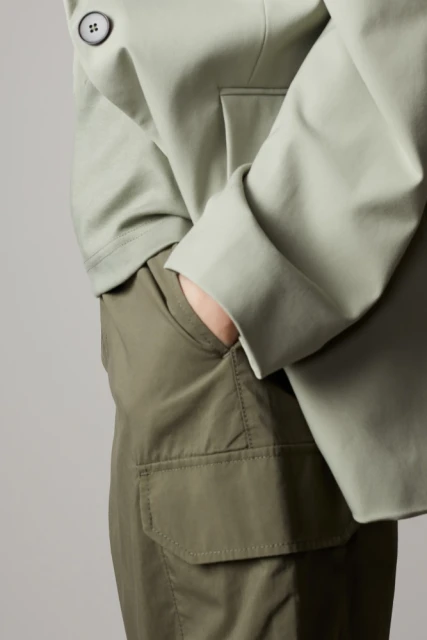 Cropped view of a person in olive green cargo pants and a light green jacket with a large black button against a neutral background.