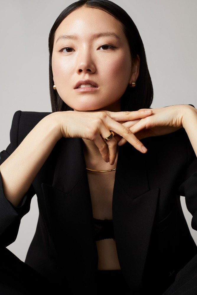 Portrait of an Asian adult female model in a black blazer and visible gold jewelry, posing in a photo studio with soft lighting.