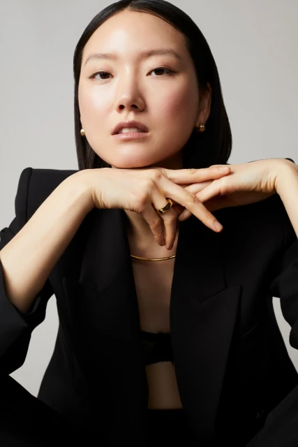 Portrait of an Asian adult female model in a black blazer and visible gold jewelry, posing in a photo studio with soft lighting.