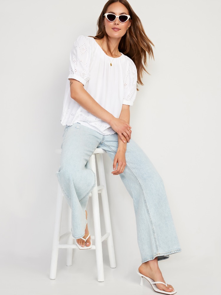Adult female wearing white top, light blue jeans, sunglasses and white heeled sandals, sitting on a white stool in a studio.