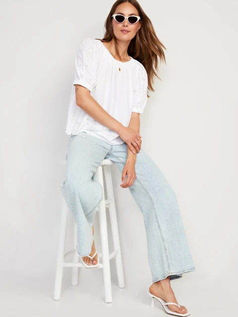Adult female wearing white top, light blue jeans, sunglasses and white heeled sandals, sitting on a white stool in a studio.