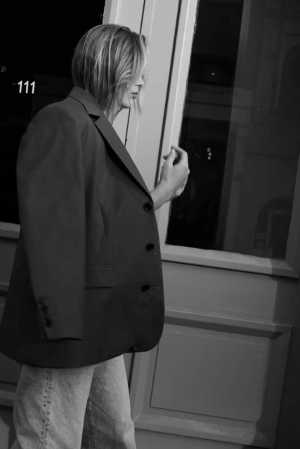 Woman in an oversized blazer and jeans walks past a building at night, the scene captured in black and white with soft focus.