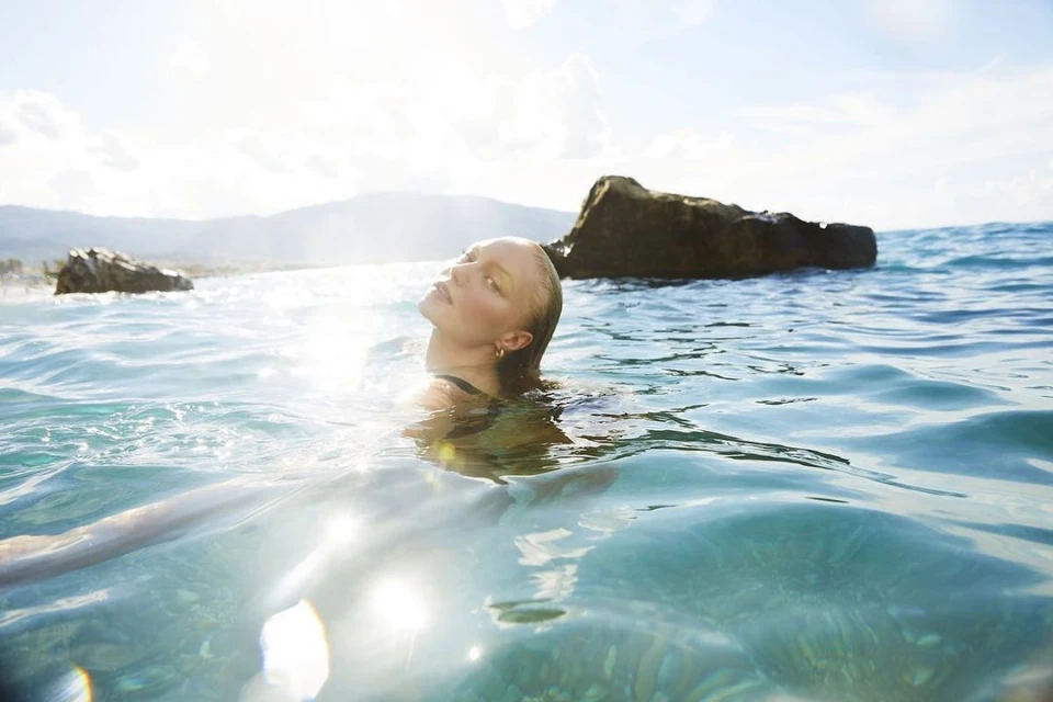Woman in the ocean with head tilted back, soaking up the sun; landscape of mountains and rocks in the distance, bright sunny day.