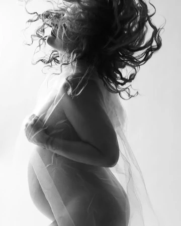 Monochrome portrait of a pregnant woman with curly hair, wrapped in a sheer white fabric, against a light background. Studio shot.