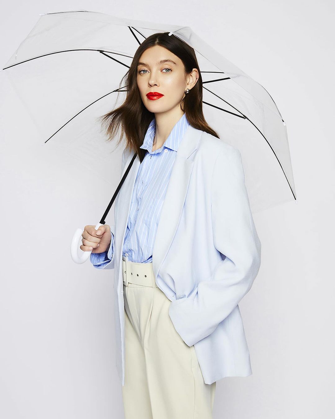 Portrait of an adult woman in a light blue blazer, light-colored pants, and red lipstick, holding a clear umbrella in a white studio.