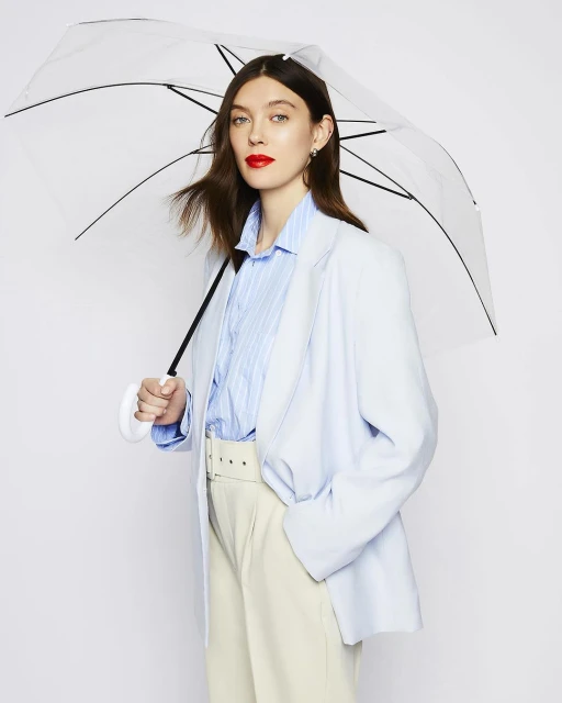 Portrait of an adult woman in a light blue blazer, light-colored pants, and red lipstick, holding a clear umbrella in a white studio.