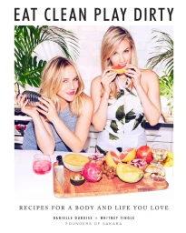 Two blonde women posing behind a wooden cutting board filled with fresh tropical fruit and a cocktail shaker.