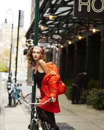 Woman in orange puffer jacket and red skirt posing with a bicycle on an urban sidewalk
