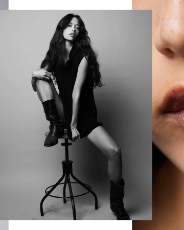 Black and white portrait of a woman with long dark hair perched on a metal stool wearing black boots.