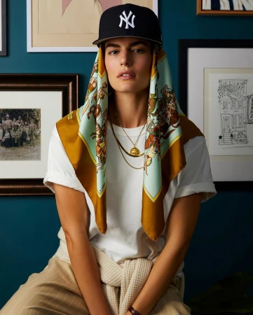 Model wearing a New York Yankees baseball cap over a patterned silk scarf, white t-shirt, and layered gold necklaces against teal wall