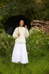 Smiling woman in a cream cable-knit sweater holding a black umbrella standing in a lush green woodland by logs.