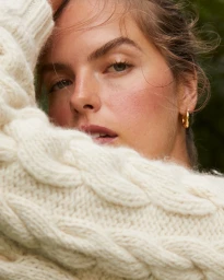 Close-up portrait of a woman wearing a cream cable-knit wool sweater and small gold hoop earrings with soft natural light.