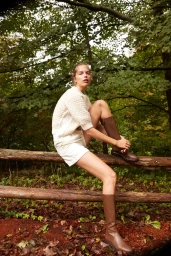 Woman in cream sweater and white shorts sitting on a wooden fence in a lush forest wearing brown boots