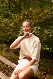 Smiling woman in a cream knit sweater and white shorts sitting on a rustic wooden railing outdoors
