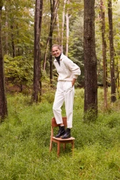 Woman in cream cable-knit sweater and white trousers standing on wooden chair in forest