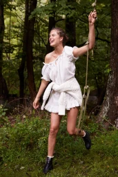 Smiling woman in a white off-the-shoulder dress holding a rope swing in a lush forest by photographer Caitlin Mitchell