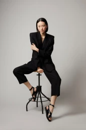 Woman with short black hair seated on a stool wearing a black blazer and trousers against a grey backdrop