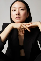 Portrait of an East Asian woman wearing a black blazer and gold jewellery against a light grey background by Caitlin Mitchell