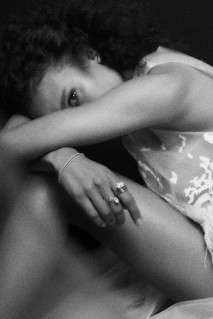 Close-up black and white portrait of a woman with curly hair resting her head on her arm, displaying stacked rings and a thin bracelet.