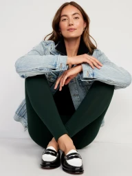 A woman seated on a white floor wearing a light blue denim jacket, black top, dark green leggings, and black and white loafers.
