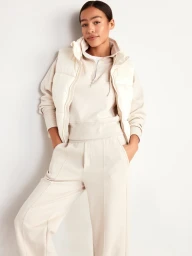 Woman in cream zip sweatshirt and puffer vest leaning against white wall