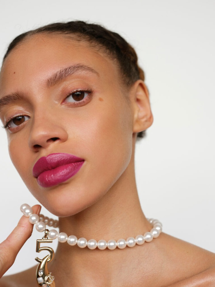 Close-up shot of an adult female model with pink lipstick and a pearl necklace with a number '5' charm, set against a plain white backdrop.