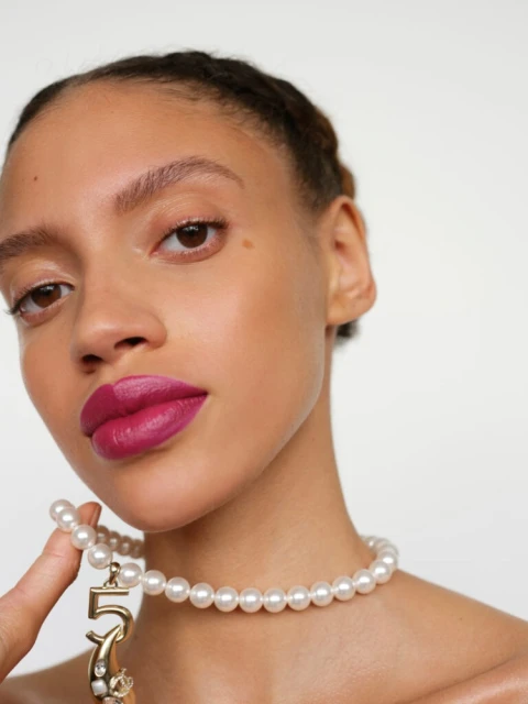 Close-up shot of an adult female model with pink lipstick and a pearl necklace with a number '5' charm, set against a plain white backdrop.