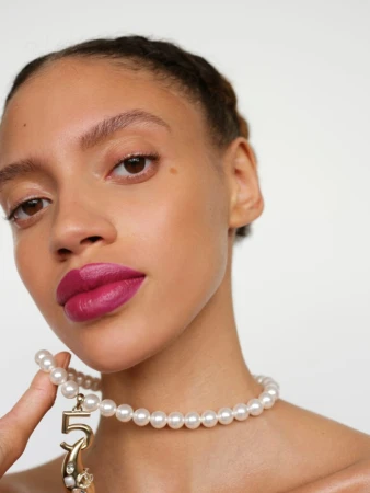 Close-up shot of an adult female model with pink lipstick and a pearl necklace with a number '5' charm, set against a plain white backdrop.