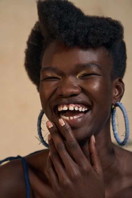 Close-up of a smiling Black woman with an afro, gold eyeshadow, and blue hoop earrings. Her hand is near her face.
