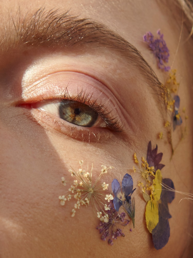 Extreme close-up of a person's green-grey eye adorned with delicate pressed purple and yellow wildflowers along the temple.
