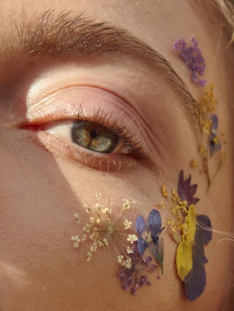 Extreme close-up of a person's green-grey eye adorned with delicate pressed purple and yellow wildflowers along the temple.