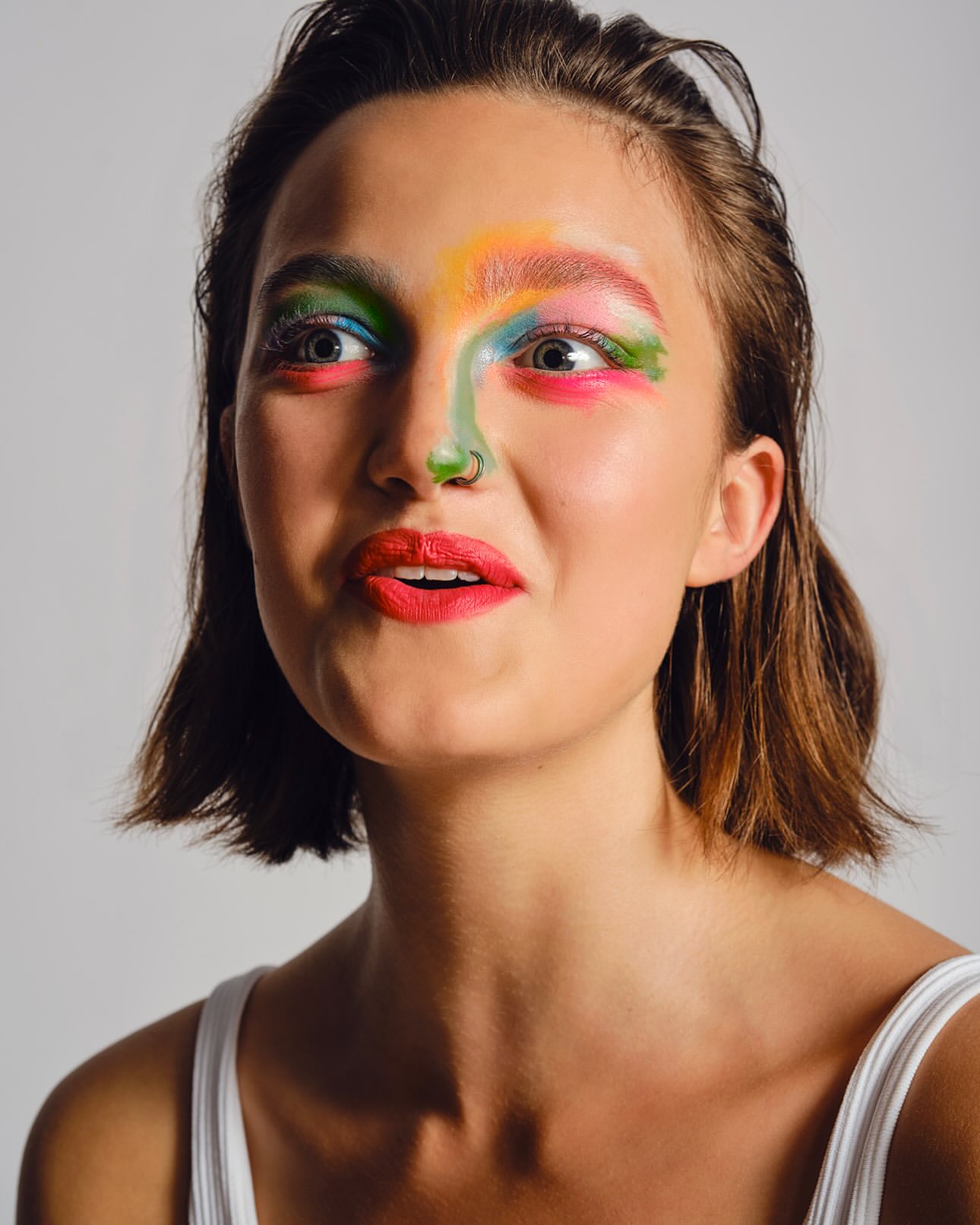 Close-up portrait of a young adult female with colorful face paint and a nose ring, set against a plain grey background in a photo studio.