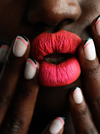 Close-up of bright pink lips and manicured hands with white nails, red lines, and dark green tips by Louise Samuelsen