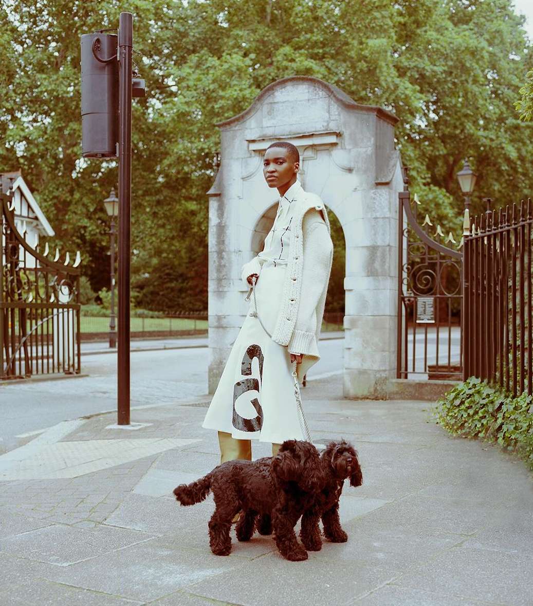 Woman in a jacket and skirt walks two black dogs on a paved walkway near a stone gate and trees in an outdoor setting.