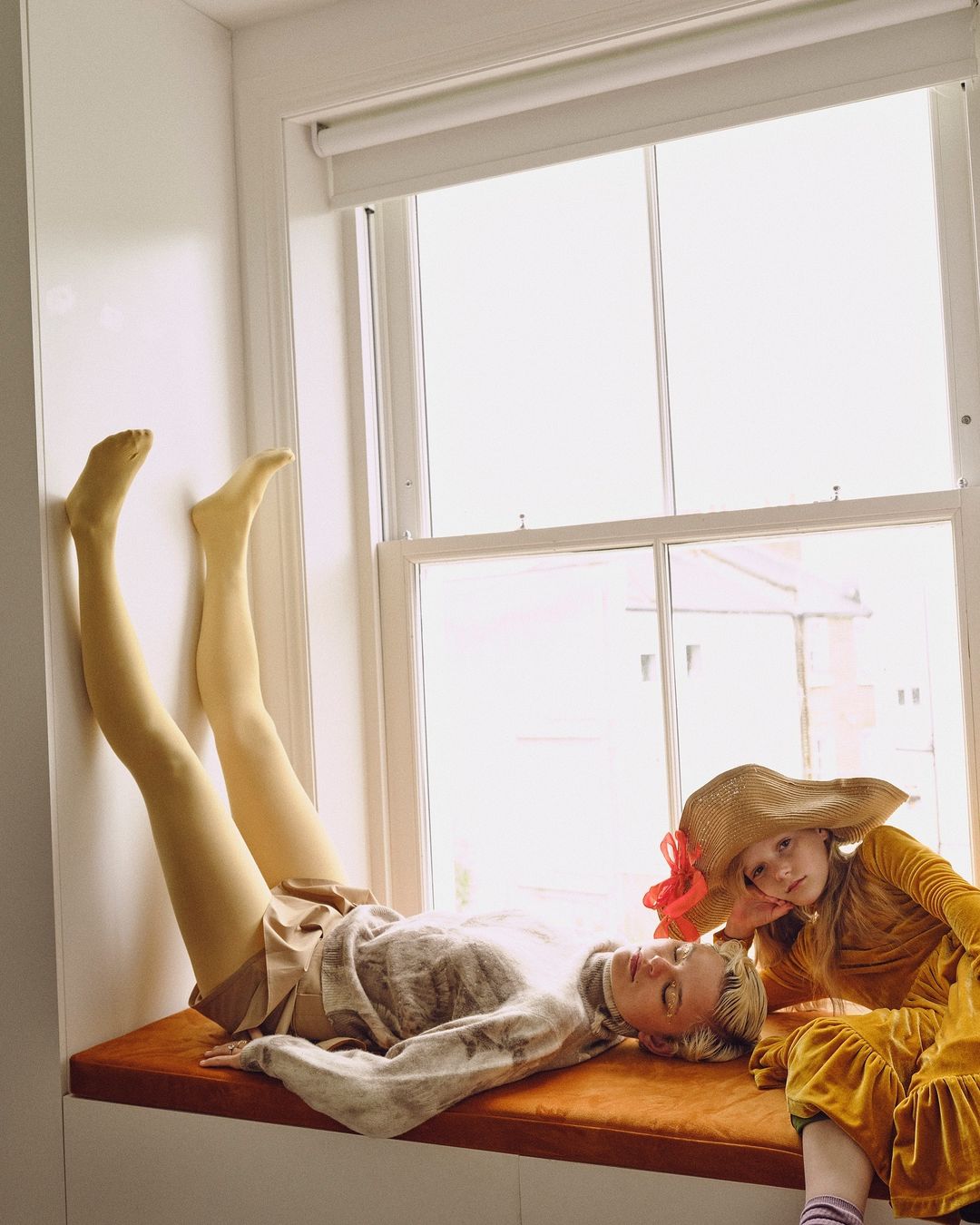 Woman and child pose near window in natural light. Adult female reclines on window seat, while young girl in sunhat sits beside.