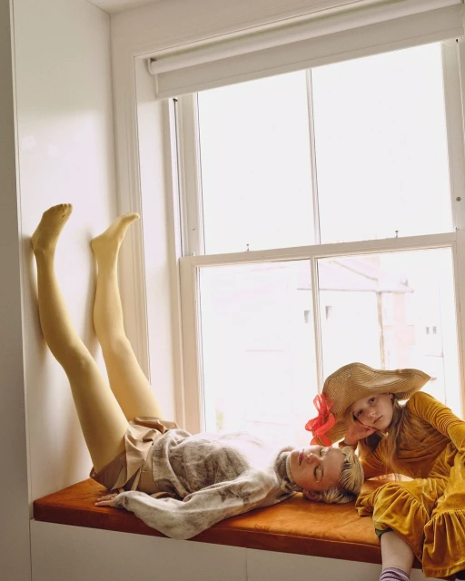 Woman and child pose near window in natural light. Adult female reclines on window seat, while young girl in sunhat sits beside.