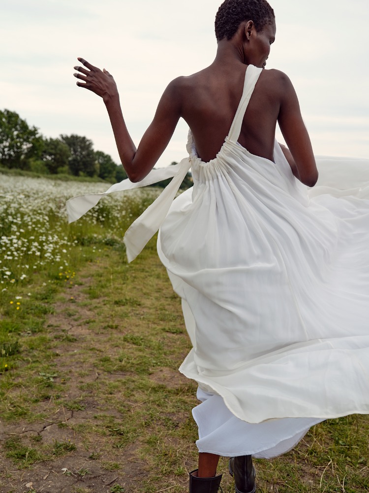 Woman in a white flowing dress walks through a field of daisies on a cloudy day, captured in an eye-level shot, showcasing nature and fashion.
