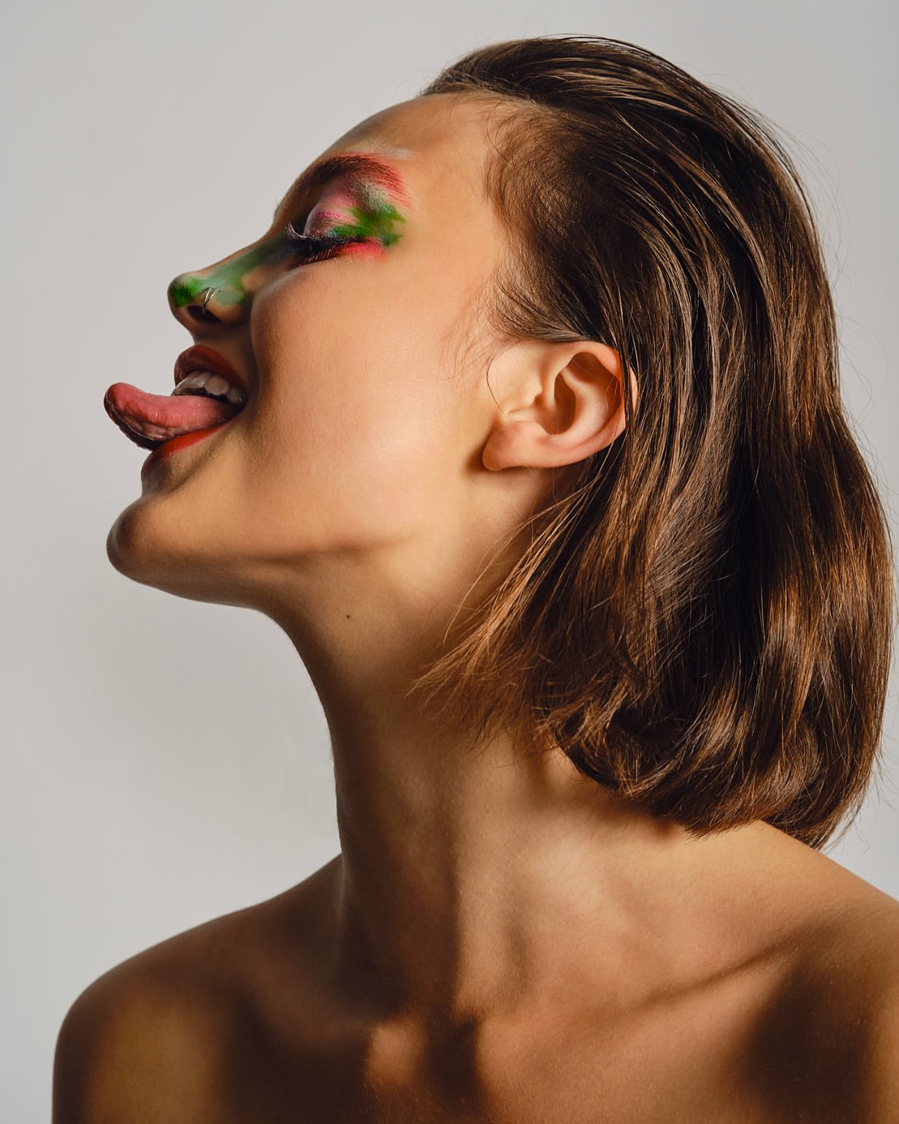 A model with creative make-up poses in a studio environment, sticking out their tongue and looking away from the camera.