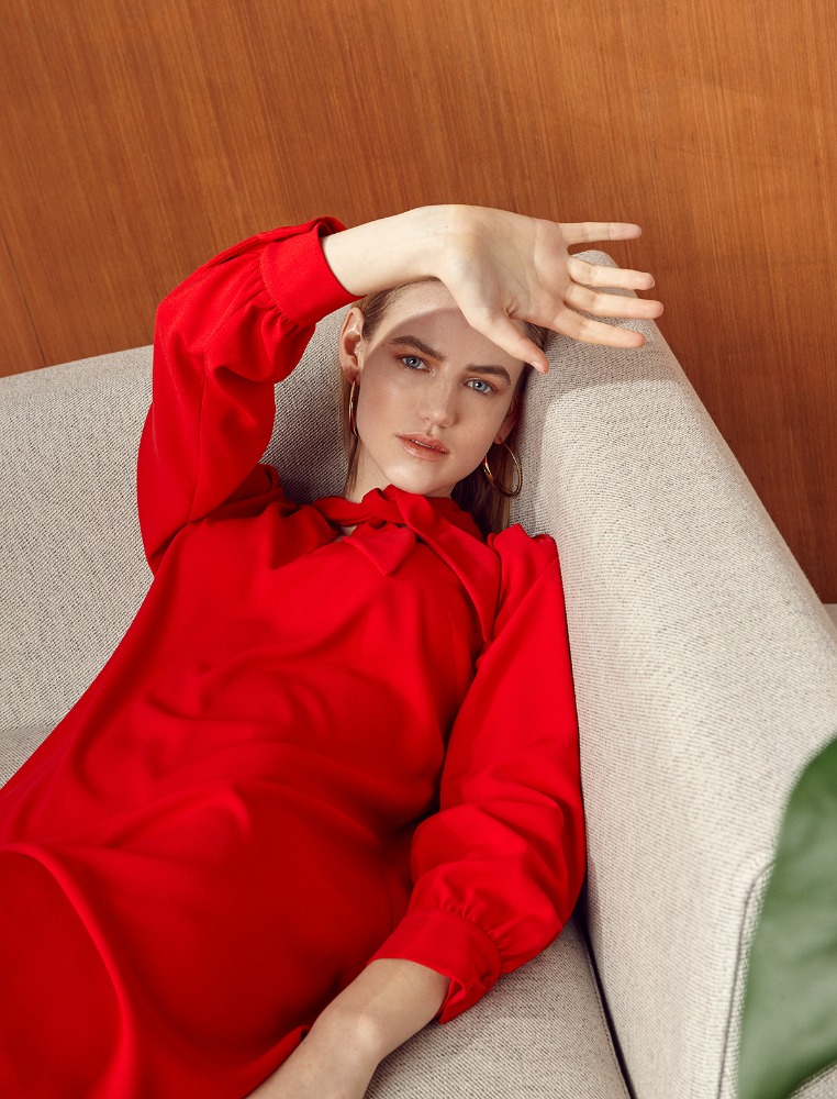 Woman in a bright red long-sleeve dress reclining on a grey couch, holding her hand up to shade her face.