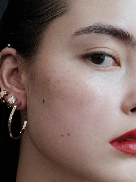 Close up of a young adult woman's face featuring gold hoop earrings, an ear piercing, fair skin, and red lipstick on her lips.