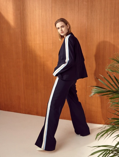 Elegant woman poses in a navy blue pantsuit with white side stripes against a warm wood paneled wall with tropical plant accents.