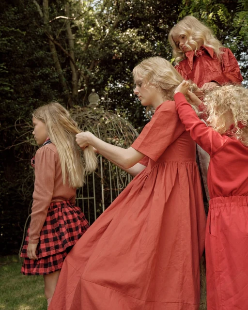 Three children and an adult woman in a yard, all dressed in red and pink clothing, are interacting with each other. A girl is having her hair braided.