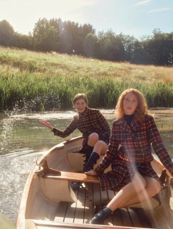 Two women wearing plaid coats seated inside a small wooden rowboat on a tranquil river with grassy banks.