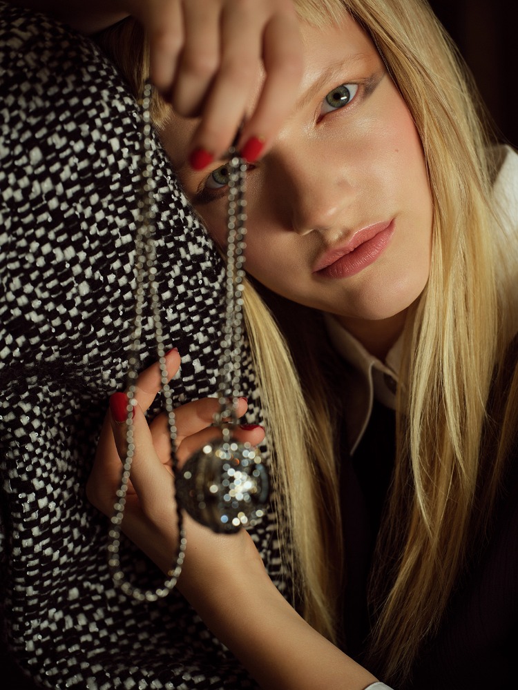 Close-up of a blond woman holding a beaded necklace with a spherical pendant, featuring a black and white patterned textile in the background.